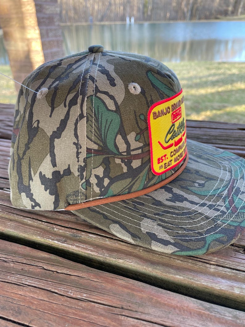 May include: A camouflage baseball cap with a brown leather band and a yellow patch that reads "Banjo Brand Music Co. Est. Eat More Catfish".