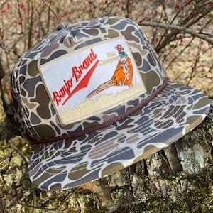 Może przedstawiać: Czapka z kamuflażowym wzorem i brązowym sznurkiem. Posiada prostokątną naszywkę z napisem "Banjo Brand" i ilustracją bażanta. Kolory to brązowy, szary i beżowy.