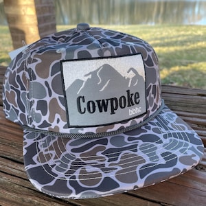 Puede incluir: Una gorra de béisbol con estampado de camuflaje y un parche gris con una cordillera y el texto "Cowpoke bbhc".