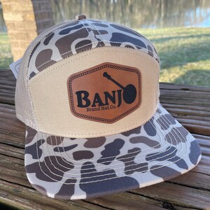Könnte beinhalten: Beige und braune Camouflage-Trucker-Mütze mit einem Lederaufnäher mit einer Banjo-Silhouette und dem Text "BANJO Brand Hat Co.". Die Mütze hat einen Mesh-Rücken und einen gebogenen Schirm.