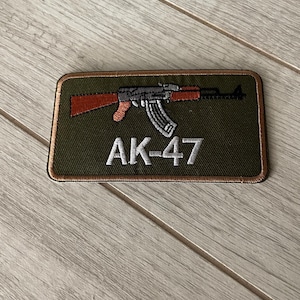 May include: An embroidered patch featuring an AK-47 rifle design. The patch has a green background with a brown and black rifle graphic and the text "AK-47" in white. The patch is rectangular with a brown border.
