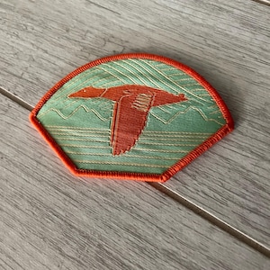 May include: Embroidered patch with an orange bird in flight on a light green background with mountain silhouettes. The patch is fan-shaped with an orange border and gold accents.