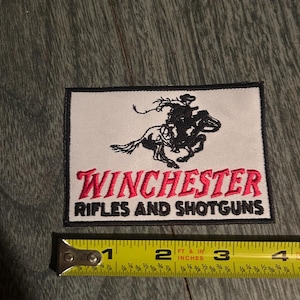 May include: A rectangular embroidered patch with a white background and black border. It features a black silhouette of a cowboy on a horse and the red text WINCHESTER above the black text RIFLES AND SHOTGUNS. A yellow measuring tape shows a width of 10.16 cm.
