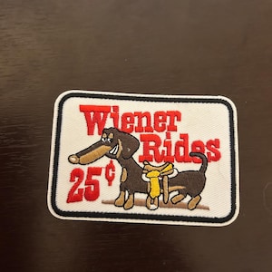May include: Embroidered patch with a white border featuring a cartoon dachshund with a yellow scarf. The text "Wiener Rides" is in red, and the price "25¢" is also displayed in red.