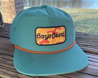 May include: A green baseball cap with an orange rope detail and a flat brim. The cap features a rectangular patch with the text "Banjo Brand" in orange and white, set against a camouflage pattern. The cap is made of a breathable material.