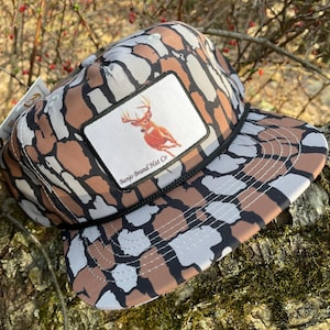 Pode incluir: Um chapéu de caminhoneiro de camuflagem marrom, cinza e preto com um remendo branco com um veado correndo e o texto "Banjo Brand Hat Co."