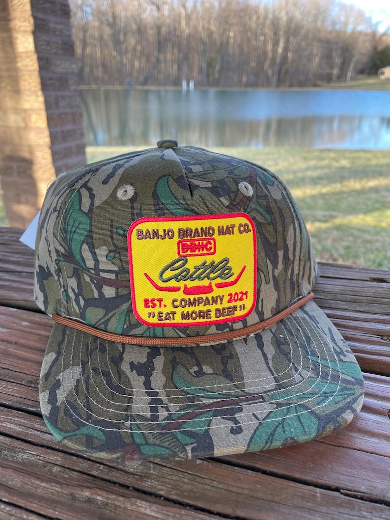 May include: A camouflage baseball cap with a brown leather band. The front of the cap features a yellow patch with red text that reads "Banjo Brand Hat Co. Cattle Est. Company 2021 "Eat More Beef!"