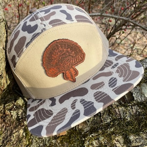 Puede incluir: Una gorra de camionero de camuflaje marrón y beige con un parche de cuero marrón que dice "OnX Hunt" en una fuente estilizada.