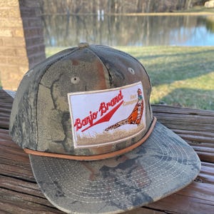 Op de afbeelding: Een camouflage baseballpet met een bruin leren bandje en een patch met de tekst "Banjo Brand Hat Company" met een illustratie van een fazant.