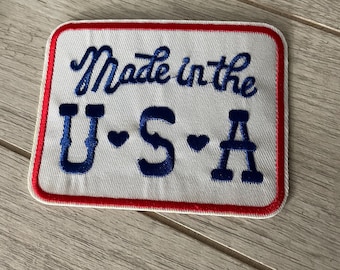 以下が含まれることがあります： 青い筆記体とブロック体の文字で「Made in the U.S.A.」と書かれた刺繍パッチ。パッチは白い背景に赤い縁取りがされています。
