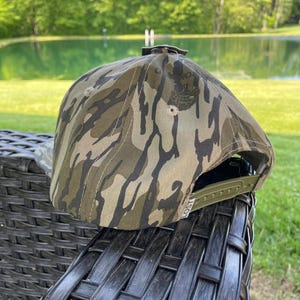 May include: A camouflage baseball cap with a green adjustable strap. The cap features a pattern of brown, green, and black shapes. The brim is curved, and the cap is sitting on a black wicker chair.