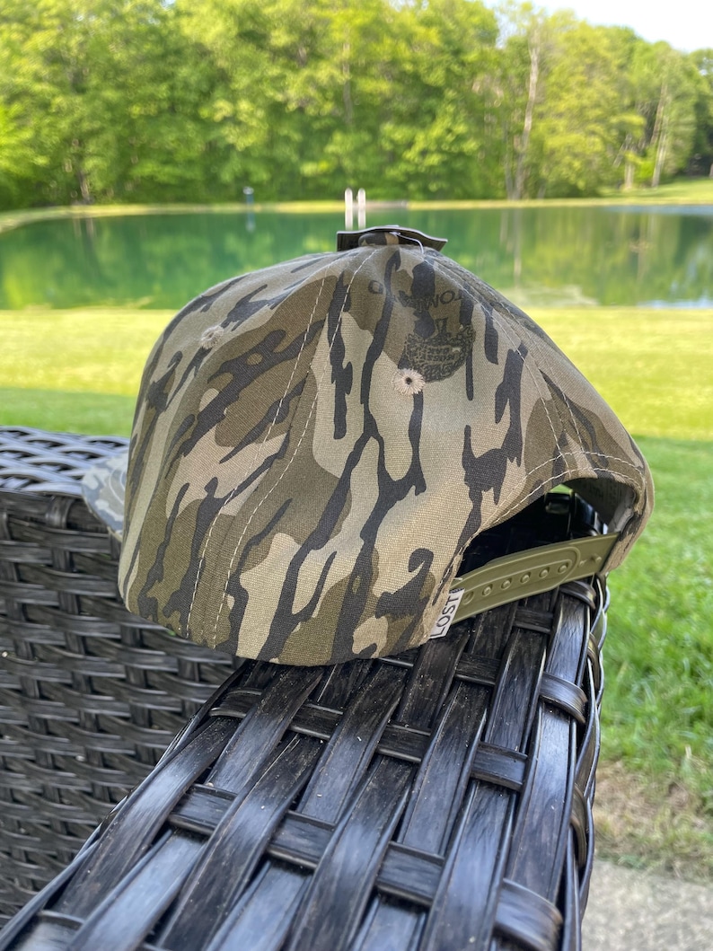 May include: A camouflage baseball cap with a green adjustable strap. The cap features a pattern of brown, green, and black shapes. The brim is curved, and the cap is sitting on a black wicker chair.