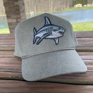 May include: A green canvas baseball cap with a white shark embroidered on the front. The cap has a grey brim and a grey strap.