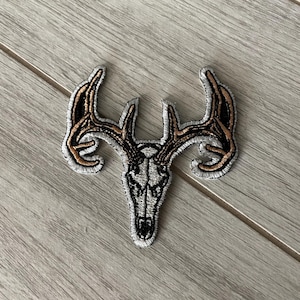 Könnte beinhalten: Gestickter Aufnäher mit einem Hirschschädel mit Geweih. Der Aufnäher hat ein detailliertes Design mit schwarzem, weißem und braunem Garn. Das Geweih hat eine braune Umrandung mit goldener Füllung. Der Schädel ist weiß mit schwarzen Details.