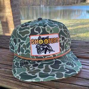 Könnte beinhalten: Eine grüne Camouflage-Trucker-Kappe mit einem braunen Seildetail und einem Patch mit der Aufschrift "SHOOTERS" in orangefarbenen und gelben Buchstaben. Der Patch zeigt eine Schwarzweißillustration einer Person, die ein Gewehr hält.