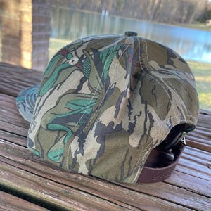May include: A camouflage patterned baseball cap with a brown leather strap. The cap has a green, brown, and white camouflage pattern.