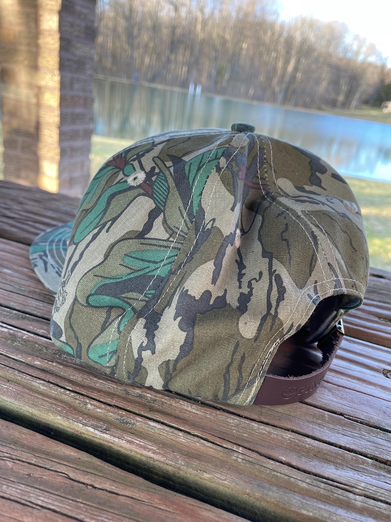 May include: A camouflage patterned baseball cap with a brown leather strap. The cap has a green, brown, and white camouflage pattern.