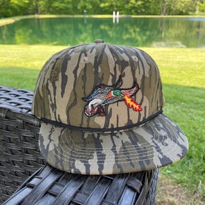 May include: Camouflage baseball cap with a duck graphic. The cap has a brown and green camouflage pattern and a black rope detail. The duck graphic is in red, green, and orange, with a flame design.