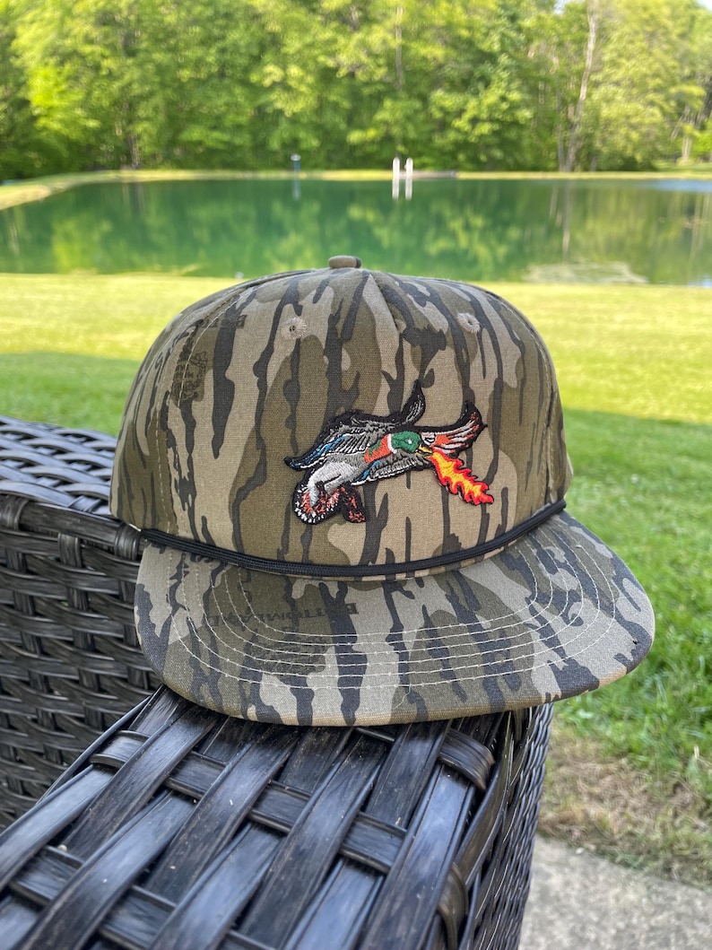 May include: Camouflage baseball cap with a duck graphic. The cap has a brown and green camouflage pattern and a black rope detail. The duck graphic is in red, green, and orange, with a flame design.