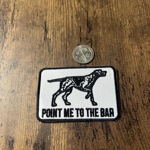 Könnte beinhalten: Ein rechteckiger Aufnäher mit schwarzem Rand und dem Text "POINT ME TO THE BAR". Der Aufnäher zeigt eine schwarz-weiße Illustration eines Hundes. Eine Münze befindet sich neben dem Aufnäher.