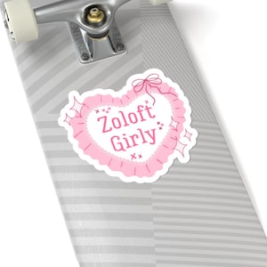 May include: A pink heart-shaped sticker with the words "Zoloft Girly" in a playful font. The sticker has a frilly border and decorative sparkles. The sticker is placed on a skateboard.