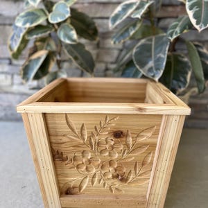 Könnte beinhalten: Rechteckiger Blumenkasten aus Holz mit einem floralen Design, das in die Frontplatte geschnitzt ist. Der Pflanzkasten ist aus hellem Holz gefertigt und hat eine natürliche Oberfläche. Die geschnitzten Blumen und Blätter verleihen dem Pflanzkasten eine dekorative Note.