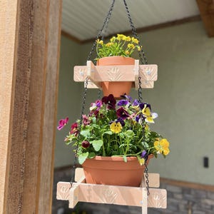 May include: Two-tiered wooden hanging planter with black metal chains. The top planter has yellow flowers and the bottom planter has purple, yellow, and white flowers.
