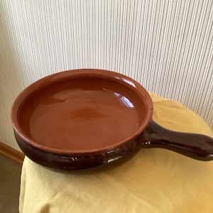 以下が含まれることがあります： 長い柄のついた茶色のセラミックフライパン。フライパンの内側は明るい茶色で、柄は濃い色合いです。フライパンは黄色の表面に置かれています。