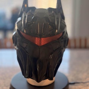 May include: A glossy black helmet featuring a pointed, bat-like silhouette with sharp, angular contours. The front includes a recessed, hexagonal-patterned visor in a metallic red finish, set within a rigid, multi-faceted shell design.