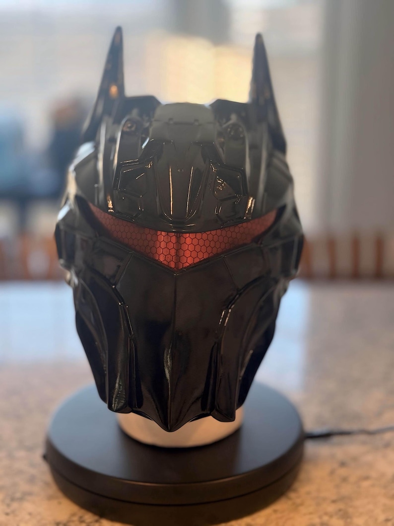 May include: A glossy black helmet featuring a pointed, bat-like silhouette with sharp, angular contours. The front includes a recessed, hexagonal-patterned visor in a metallic red finish, set within a rigid, multi-faceted shell design.