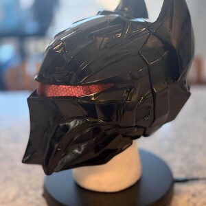 May include: A glossy black full-face helmet featuring a sharp, angular design with two prominent pointed ears at the top. The eye area displays a recessed, textured red visor with a distinct hexagonal pattern visible across the horizontal opening.