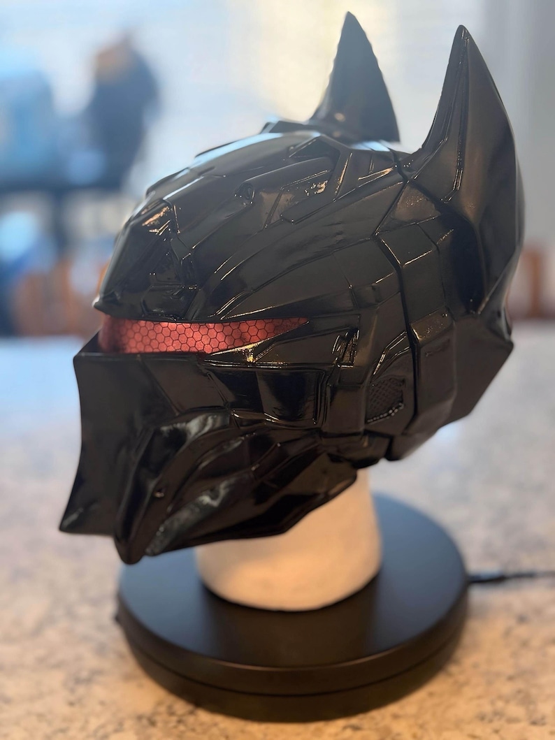 May include: A glossy black full-face helmet featuring a sharp, angular design with two prominent pointed ears at the top. The eye area displays a recessed, textured red visor with a distinct hexagonal pattern visible across the horizontal opening.