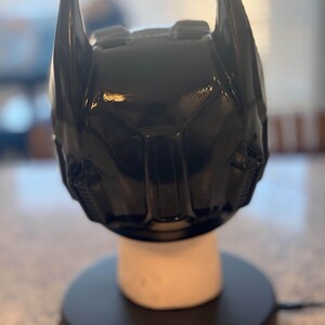 May include: A glossy black Batman cowl helmet featuring two prominent pointed ears at the top, a sculpted facial structure with defined brow ridges, and a central nose bridge, displayed on a white cylindrical stand atop a circular black base.