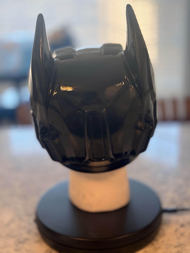 May include: A glossy black Batman cowl helmet featuring two prominent pointed ears at the top, a sculpted facial structure with defined brow ridges, and a central nose bridge, displayed on a white cylindrical stand atop a circular black base.