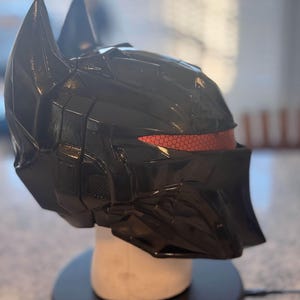 May include: A glossy black full-face helmet featuring a sharp, angular design with two prominent pointed ears at the top. The helmet includes a horizontal eye visor with a distinct red honeycomb pattern texture across the front viewing area.