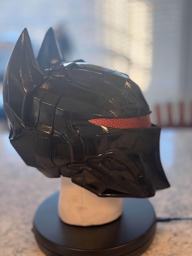 May include: A glossy black full-face helmet featuring a sharp, angular design with two prominent pointed ears at the top. The helmet includes a horizontal eye visor with a distinct red honeycomb pattern texture across the front viewing area.