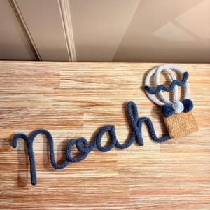 May include: A blue knitted name "Noah" with a hot air balloon design. The balloon is white and blue with a woven basket. The name is in a cursive font.