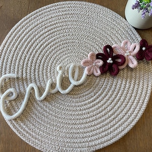 May include: A white crocheted name sign spelling "Erica" with pink and burgundy crocheted flowers.