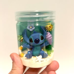 Puede incluir: Un tarro de pl&aacute;stico transparente que contiene una figura azul de Stitch sosteniendo un cono de helado. El tarro est&aacute; lleno de arena, un l&iacute;quido verde y elementos decorativos como una estrella de mar, flores y un sol. El tarro es sostenido por una mano.