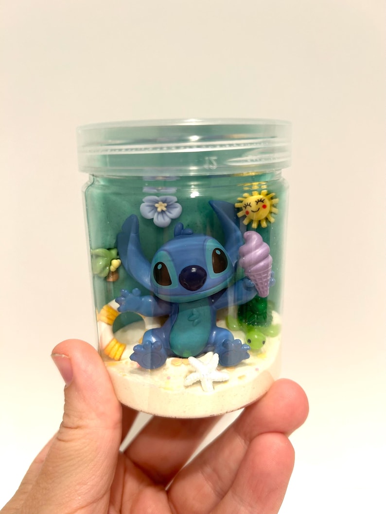 Puede incluir: Un tarro de pl&aacute;stico transparente que contiene una figura azul de Stitch sosteniendo un cono de helado. El tarro est&aacute; lleno de arena, un l&iacute;quido verde y elementos decorativos como una estrella de mar, flores y un sol. El tarro es sostenido por una mano.