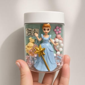 May include: A clear plastic jar with a white lid containing a small figurine of a character in a blue dress holding a gold star wand. Inside are miniature items including a book labeled Once Upon A Time, a yellow castle, a pink mirror, and crystals.