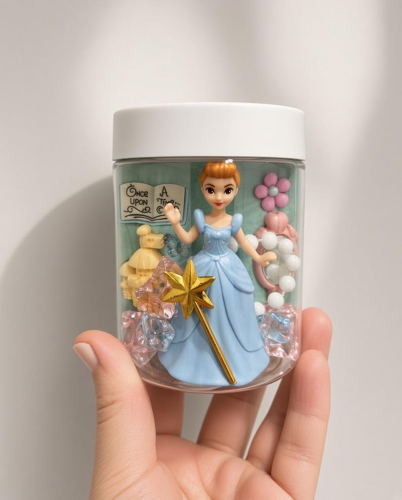 May include: A clear plastic jar with a white lid containing a small figurine of a character in a blue dress holding a gold star wand. Inside are miniature items including a book labeled Once Upon A Time, a yellow castle, a pink mirror, and crystals.