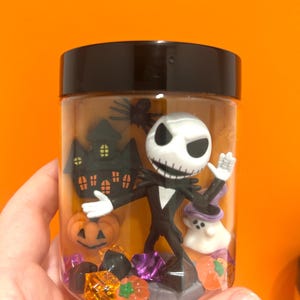May include: A clear plastic jar with a black lid filled with Halloween-themed figurines. The jar contains a Jack Skellington figure, a haunted house, a pumpkin, and a ghost. The background is a bright orange.