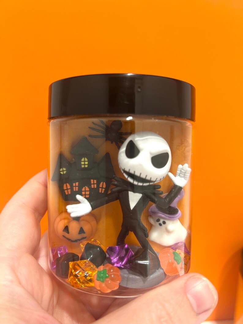 May include: A clear plastic jar with a black lid filled with Halloween-themed figurines. The jar contains a Jack Skellington figure, a haunted house, a pumpkin, and a ghost. The background is a bright orange.