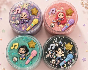 May include: Four circular containers filled with colorful slime, each topped with a miniature figurine of a pop star, a microphone, a musical note, a gold glitter star, and a small disco ball. The slimes are purple, pink, teal, and black.