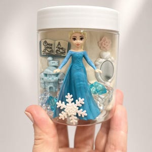 May include: A clear plastic jar with a white lid containing a small blonde figurine in a blue dress, a blue castle, a white book with the text Once Upon A Time, a white mirror, a white snowflake, a pink flower, and several blue translucent gems.