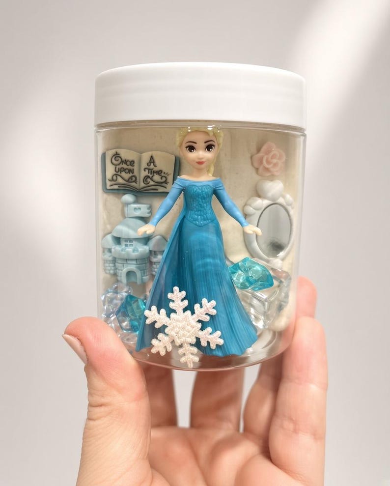 May include: A clear plastic jar with a white lid containing a small blonde figurine in a blue dress, a blue castle, a white book with the text Once Upon A Time, a white mirror, a white snowflake, a pink flower, and several blue translucent gems.