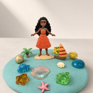 May include: A small Moana figurine stands on a sandy base atop a circular light blue clay disc. The scene includes a miniature palm tree, a small sailboat, a pink starfish, a pearlescent seashell, and several colorful translucent plastic gems.