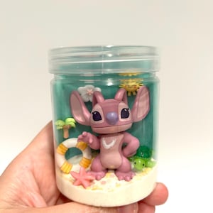 Puede incluir: Un tarro de pl&aacute;stico transparente que contiene una figura rosa de Angel de Lilo & Stitch, rodeada de decoraciones en miniatura con tem&aacute;tica de playa. El tarro incluye un peque&ntilde;o salvavidas, estrellas de mar, palmeras y una tortuga verde, todo sobre una base arenosa y un fondo turquesa.