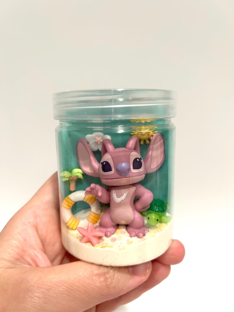 Puede incluir: Un tarro de pl&aacute;stico transparente que contiene una figura rosa de Angel de Lilo & Stitch, rodeada de decoraciones en miniatura con tem&aacute;tica de playa. El tarro incluye un peque&ntilde;o salvavidas, estrellas de mar, palmeras y una tortuga verde, todo sobre una base arenosa y un fondo turquesa.